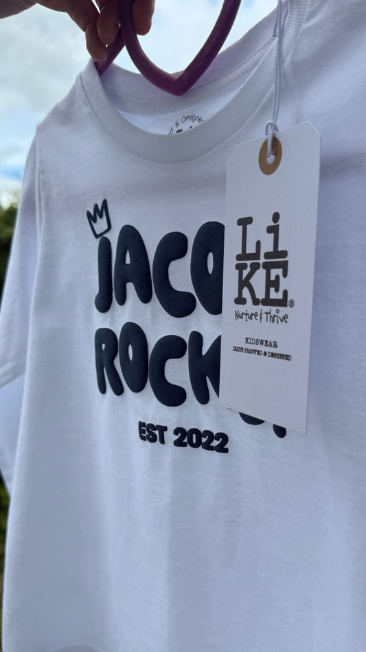 White t-shirt with 'Jacob Rocks' text and a tag, held against a blurred natural background.